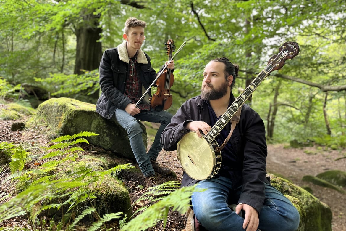 2 men sitting on rocks in a wood 1 with a violin and 1 with a banjo